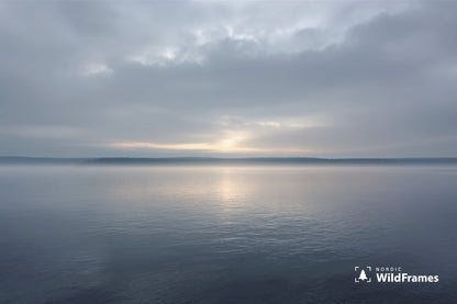 Minimal calm lake with sunlight breaking through clouds above a misty forest horizon, Finland