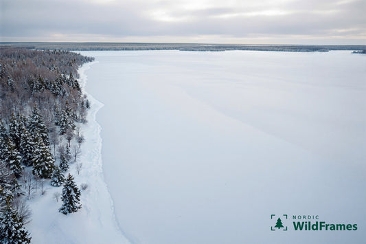 Aerial view of a frozen snow-covered lake and frosted forest shoreline under overcast winter sky, Finland